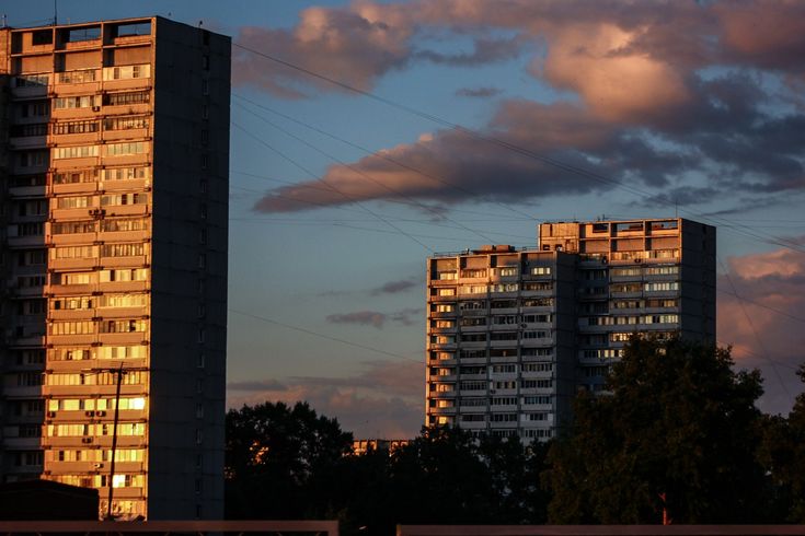 the image shows two tall apartment buildings at sunset. the building on the left is illuminated by the warm light of the setting sun, while the building on the right is in shadow. the sky is a mix of dark clouds and bright orange hues.  the overall mood is serene and slightly melancholic. green trees are visible in the foreground and between the buildings.  the image likely depicts a city in eastern europe, possibly russia, given the style of architecture.  power lines are visible across the sky.