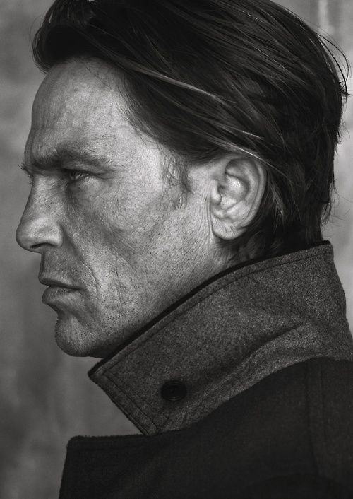 a black and white portrait of a mature man in profile. the man has weathered skin and noticeable wrinkles, suggesting age. he is wearing a coat with a button visible near his collar. the image captures the details of his face, including his hair and ear. the texture of the coat and the man's skin are prominent.
