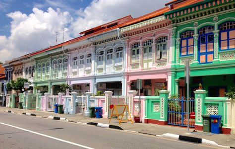 the image showcases a row of vibrantly colored shophouses in singapore.  these traditional buildings, characteristic of peranakan architecture, are painted in a range of pastel shades, including pinks, light blues, greens, and yellows. the shophouses are neatly aligned, each featuring ornate detailing on their facades.  the street in front is clean and relatively quiet, with a few waste bins visible.  the sky is partly cloudy, and the overall scene is bright and cheerful, capturing the essence of singapore's architectural heritage and its lively urban environment.