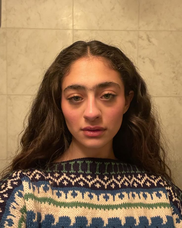 a closeup portrait of a woman with long curly brown hair, green eyes, and fair skin, wearing a fair isle sweater. she is facing the camera with a neutral expression. the background appears to be a tiled wall.