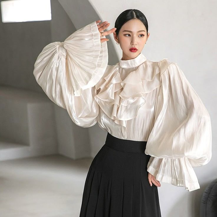 a woman is wearing an elegant white blouse with ruffles and large sleeves, paired with a black pleated skirt. she is posing with one hand raised, showcasing her jewelry. the background is a neutral color.