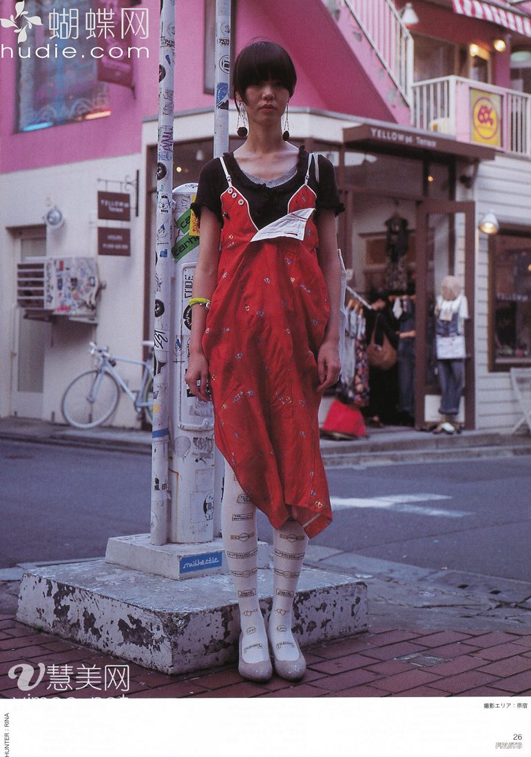 a young woman with short black hair stands on a city sidewalk in front of a pink building. she wears a long, sleeveless red dress with white patterns, white tights with a design on them, and white shoes. the image captures a street style aesthetic, likely in harajuku, japan. the lighting is natural and slightly overcast, and the perspective is from eye level.