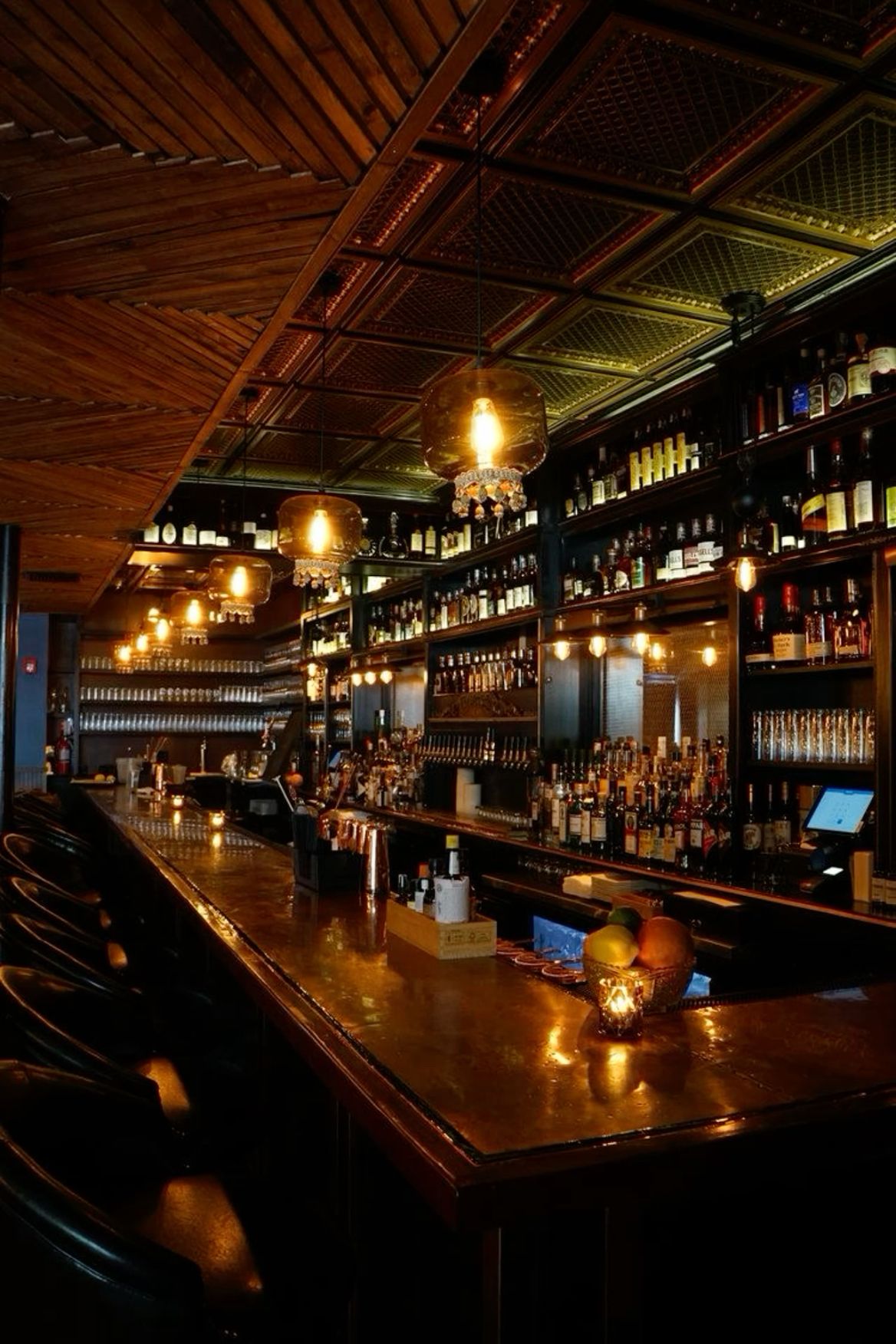 the image shows the inside of a bar, with a long copper bar counter lined with bar stools on the left and shelves full of liquor bottles in the back. the warm, dim lighting creates a cozy and inviting atmosphere.