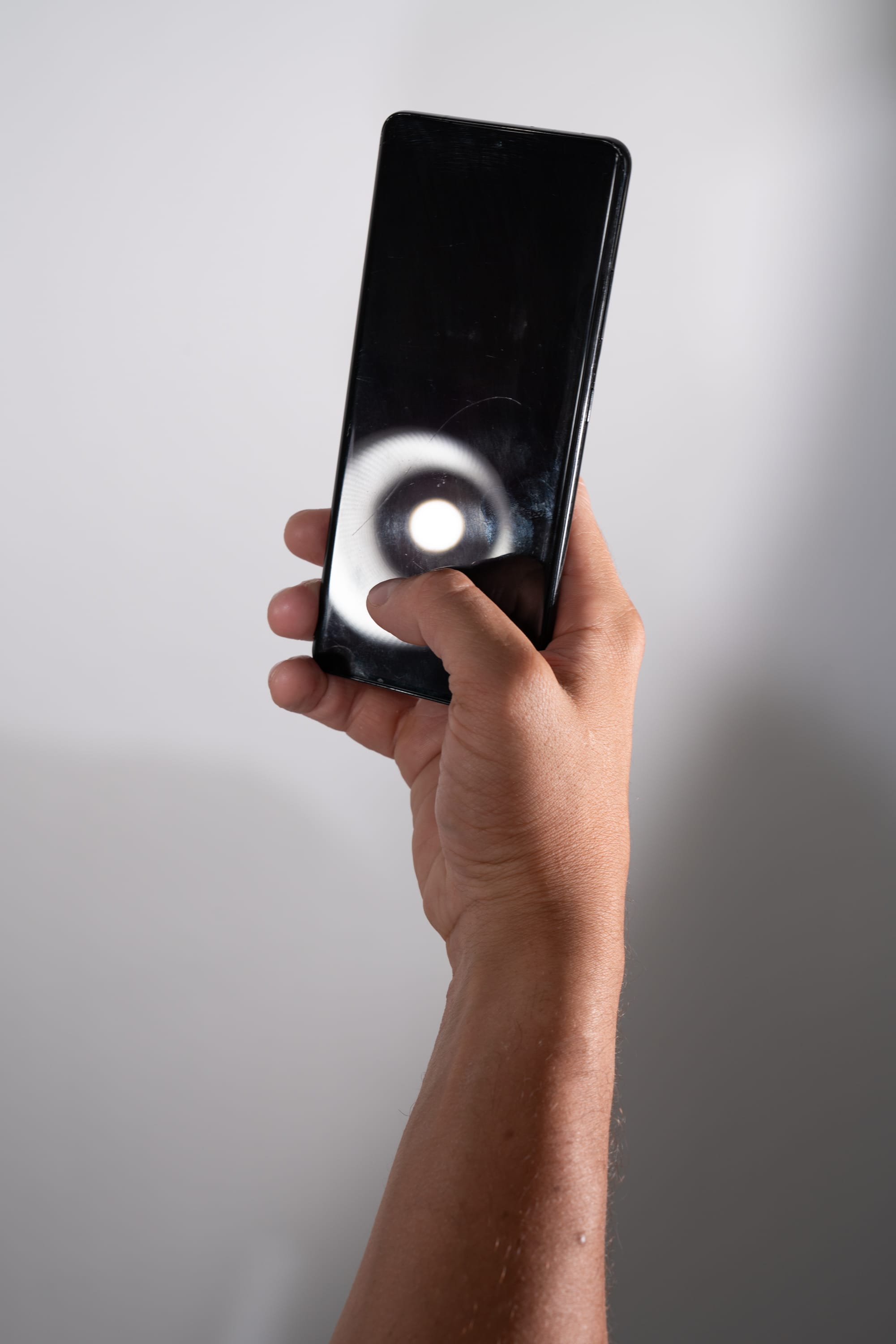a man's hand holds a smartphone up against a white background. the smartphone's screen displays a bright circle of light.