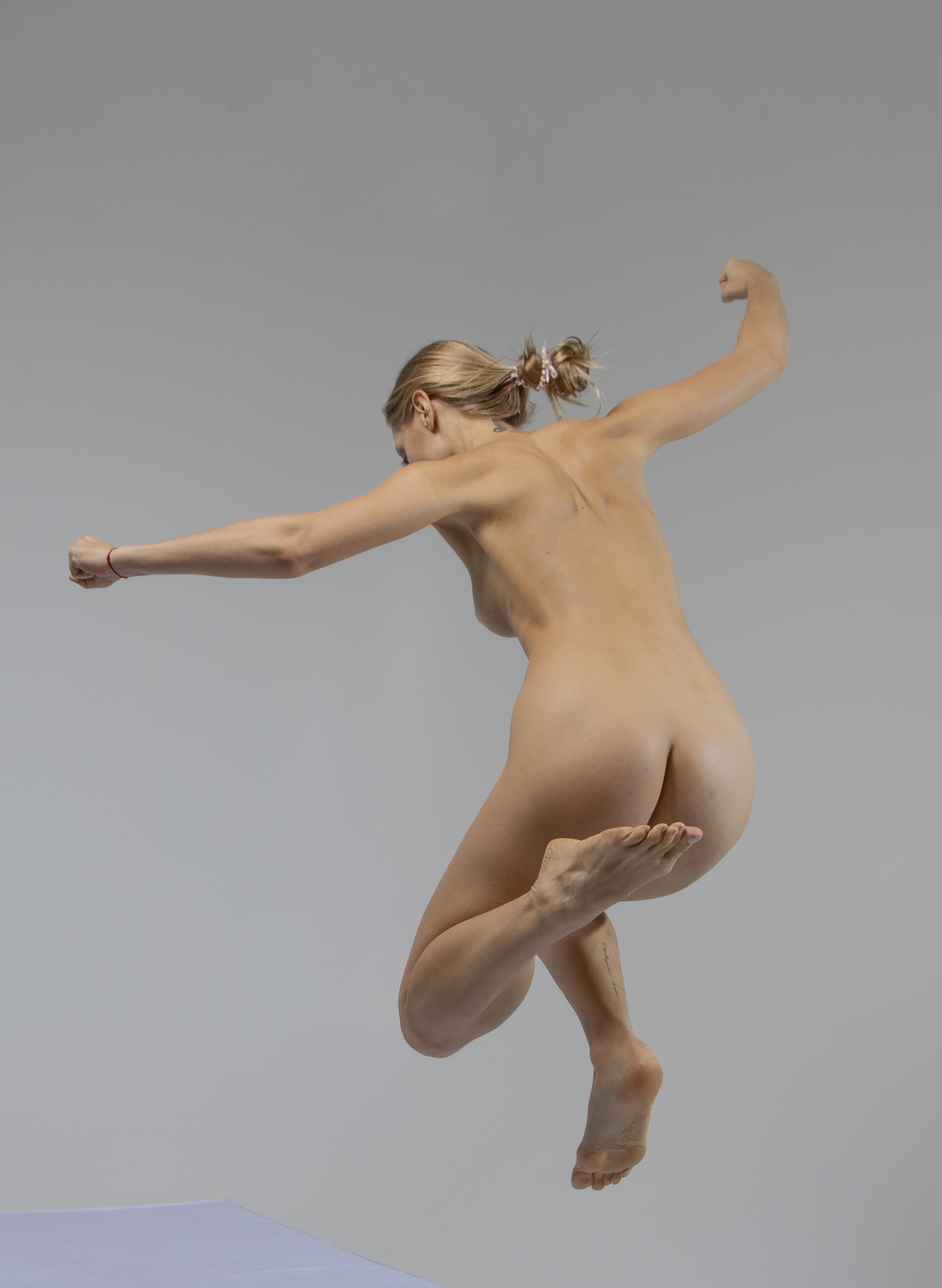 a nude blonde woman with her hair tied back is captured midjump against a neutral gray background. her body is bare and facing away from the viewer, highlighting her back, buttocks, and the muscles in her legs. her arms are outstretched, one with a fist clenched, suggesting movement or a burst of energy. she appears to be in a fitness or athletic pose, emphasizing strength and agility. a glimpse of a white surface is visible at the bottom of the frame, possibly the edge of a platform or trampoline.