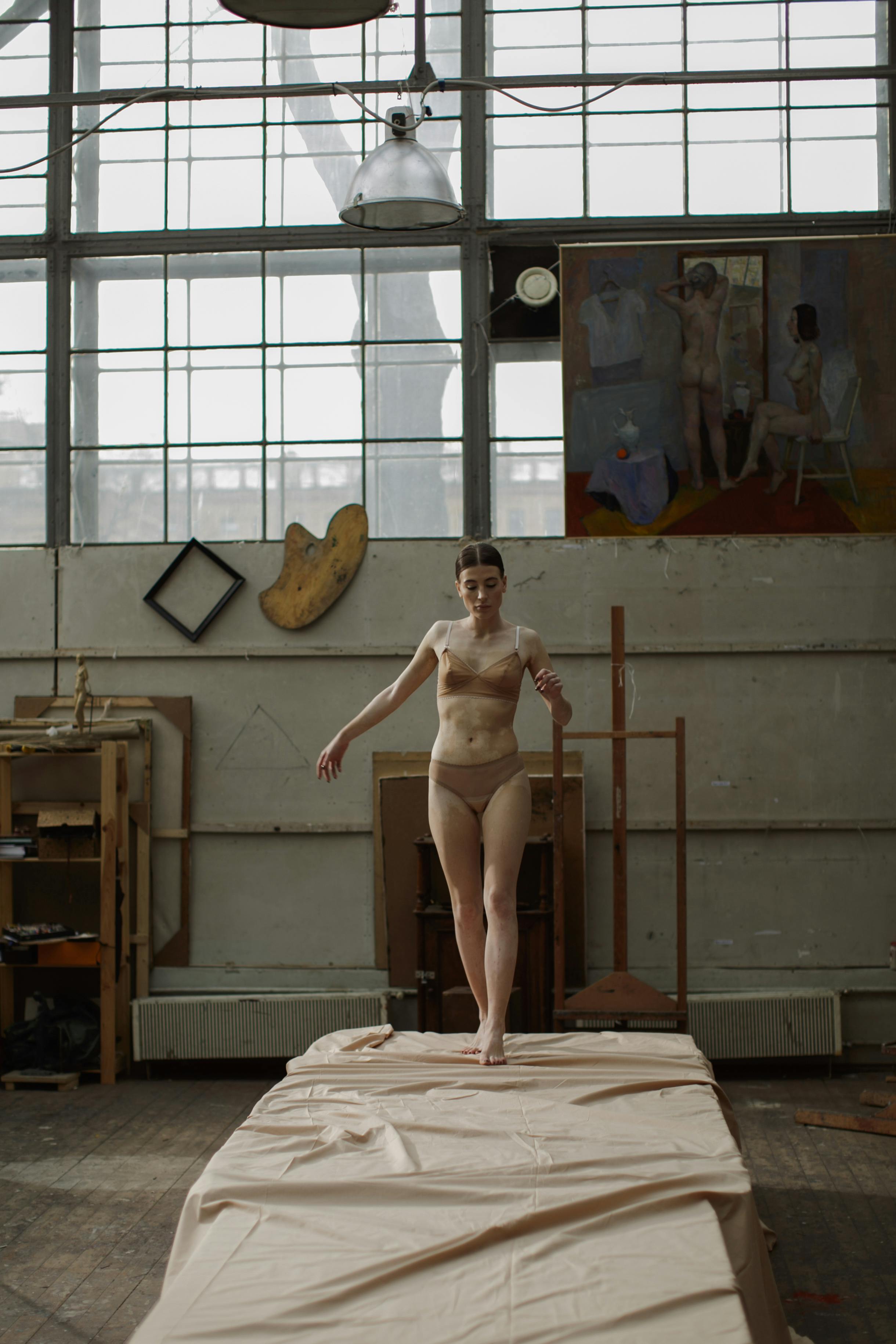 a young woman, wearing a nudecolored bra and panties, walks across a large canvas laid out on the floor of an art studio. the studio is bright with natural light coming from large windows, and features an easel, artwork, and painting supplies.  the woman's pose and the overall aesthetic of the space suggest a connection to art and the human form. the color palette is muted and neutral.