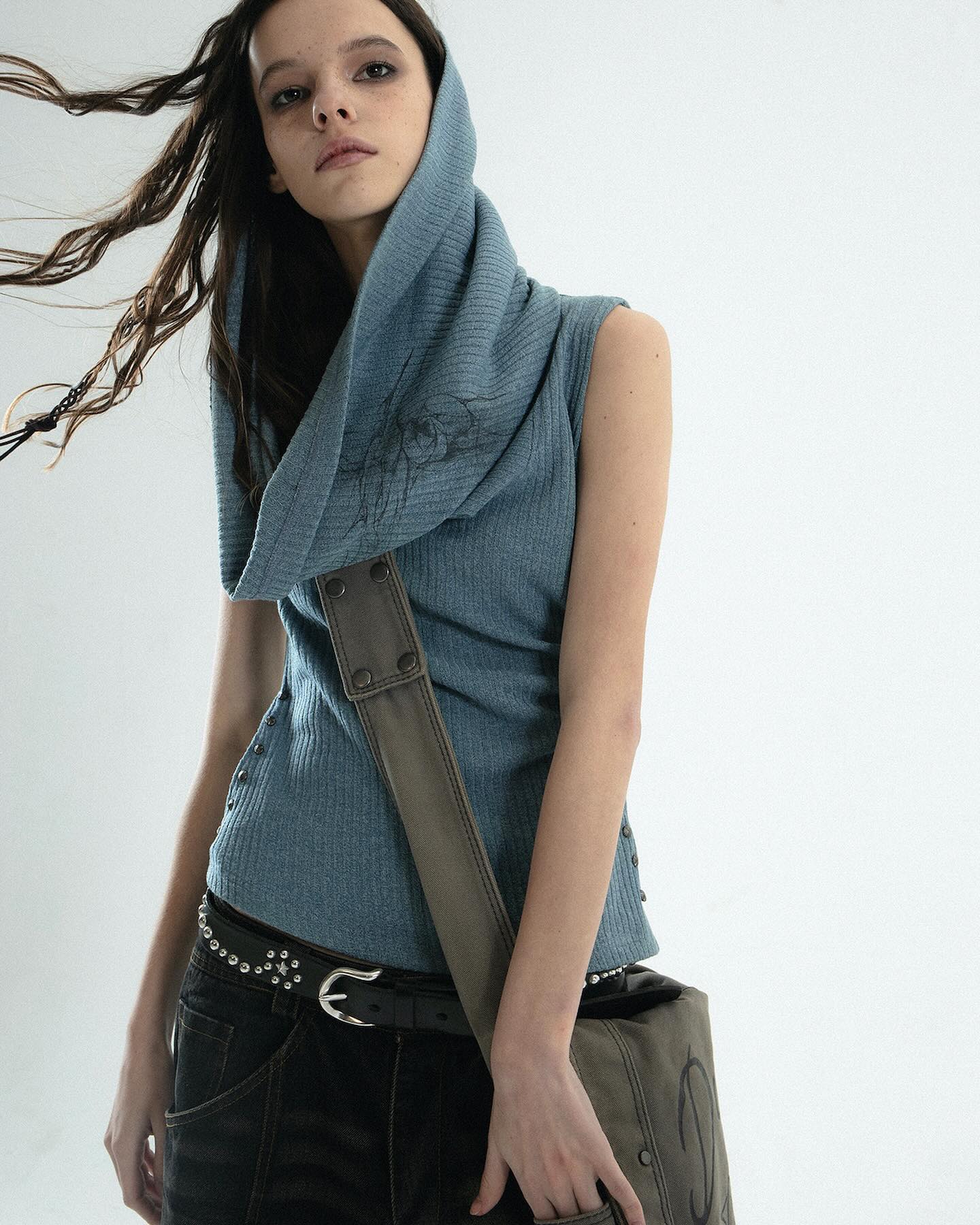 a young woman with long brown hair is modeling a casual outfit.  she is wearing a light blue ribbed knit sleeveless top with a large cowl hood. the top features subtle embroidered detailing. she's paired the top with darkwash jeans and a studded belt. a dark olive green messenger bag hangs across her body. the woman is posing against a plain white background. the overall style is relaxed and slightly edgy.