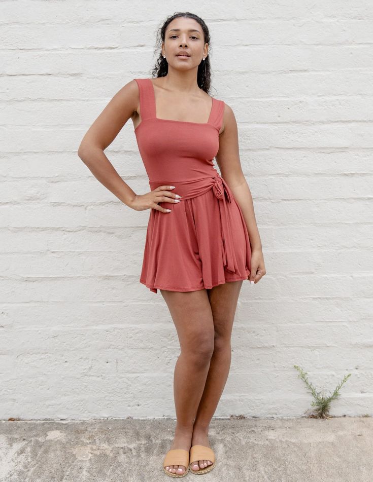a young woman with tanned skin and dark curly hair is posing in front of a white brick wall. she is wearing a burnt orange romper with a square neckline and a tie waist. she is also wearing brown sandals. the lighting is natural and bright, and the overall feel of the image is summery and stylish.