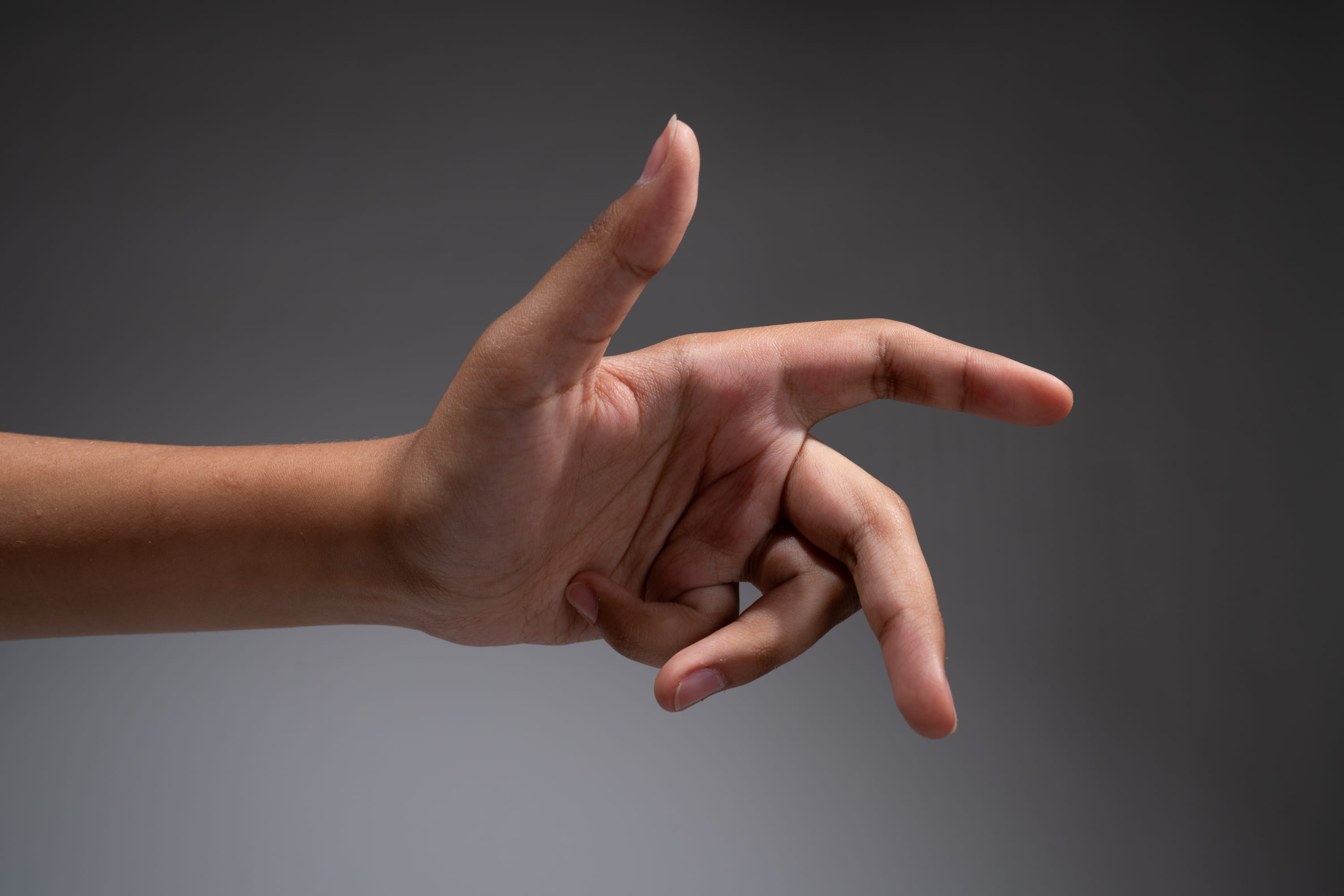 a hand is extended against a gray background. the index finger is pointing, while the other fingers are curled into the palm. the hand is positioned in a way that suggests it's directing attention to something offscreen. the lighting is soft and even, highlighting the shape and texture of the hand.