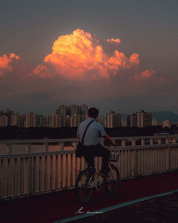 a man is riding a bicycle on a bridge over a river. the city skyline is in the background, and a large, vibrant orange and yellow cloud dominates the sky during sunset. the overall mood is serene and peaceful.