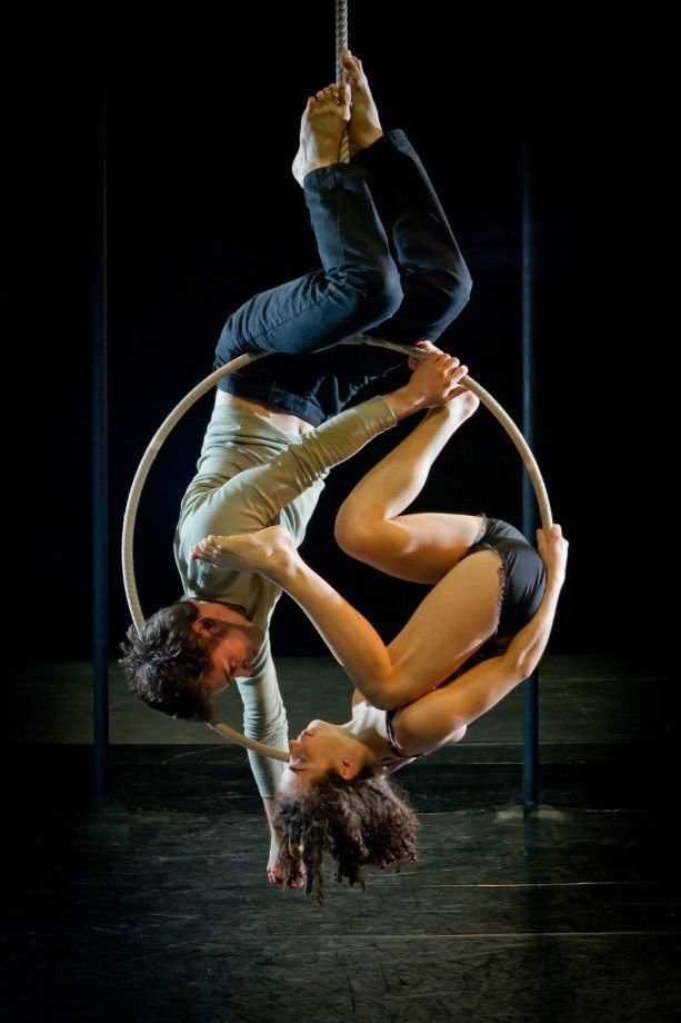 a male and female acrobat perform an aerial routine in a circus ring. they are suspended by a rope above the ring, while the male artist hangs upside down, with his legs wrapped around the ring, and the female artist is  suspended by a rope from the top of the ring, with her legs stretched out and her body bent backwards.  they are both wearing black clothing and have their bodies bent in interesting poses.  the black background accentuates their movement.  the photo was likely taken during a performance.