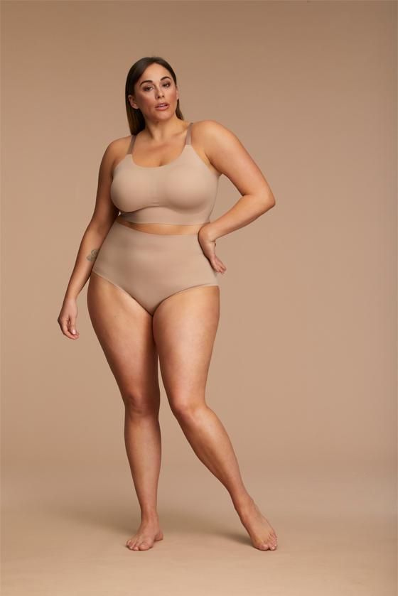 a plussize model poses confidently wearing a beige seamless bra and highwaisted underwear set. the background is a neutral, light brown tone. the image highlights body positivity and the comfortable fit of the lingerie. the model's pose and expression convey selfassurance and comfort in her own skin. the lighting is soft and even.