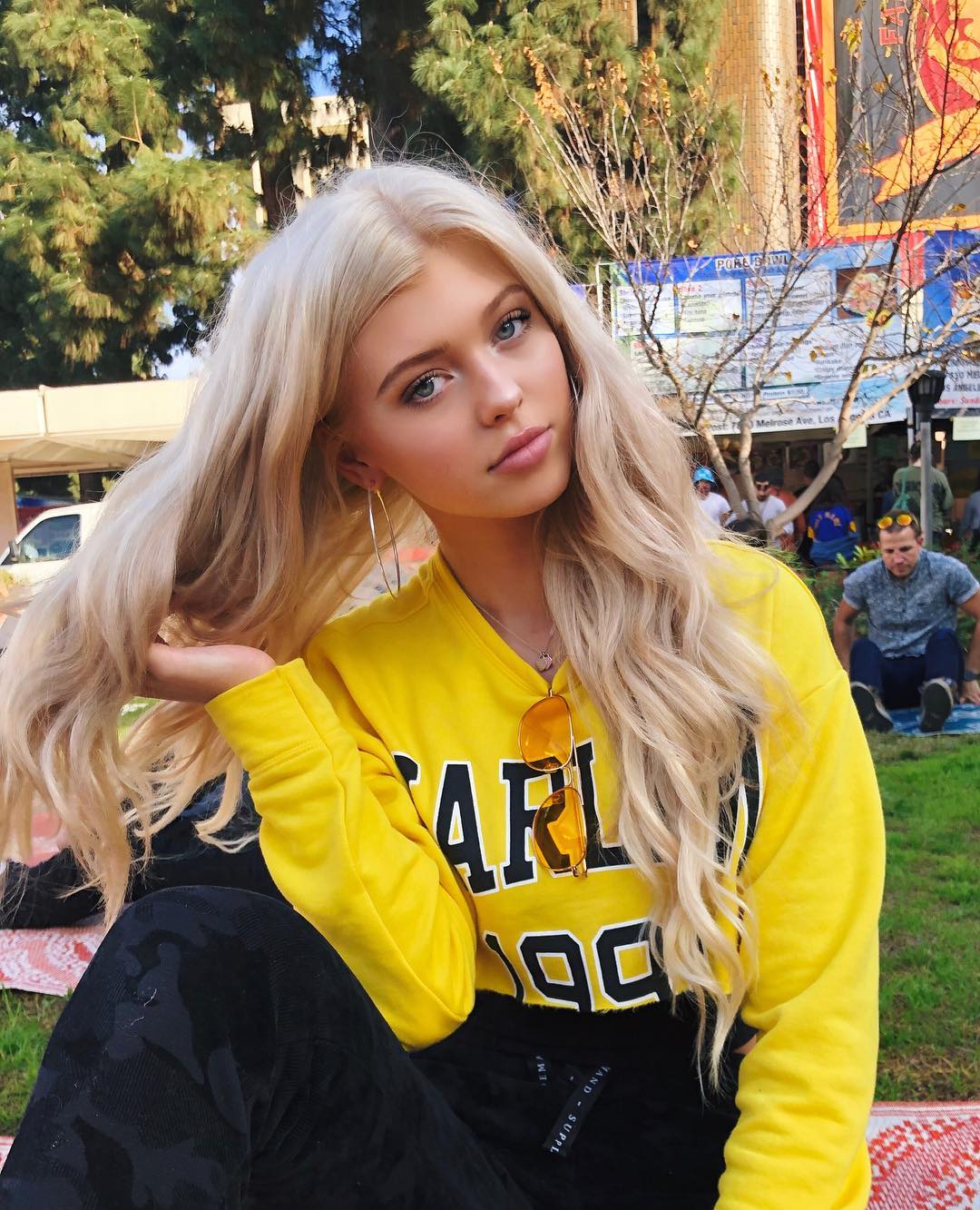 a young woman with blonde hair is sitting on the grass outdoors. she is wearing a yellow sweater with black accents, black pants, and hoop earrings. sunglasses are hanging on her sweater. there are trees and a building in the background.