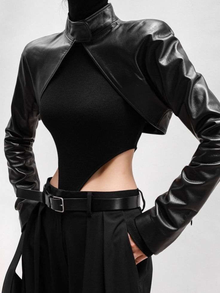 a model wearing a black cropped leather jacket, black top, black pants and a belt. the model is posed with their hand in their pocket, looking to the right. the photo has a studio shot background.