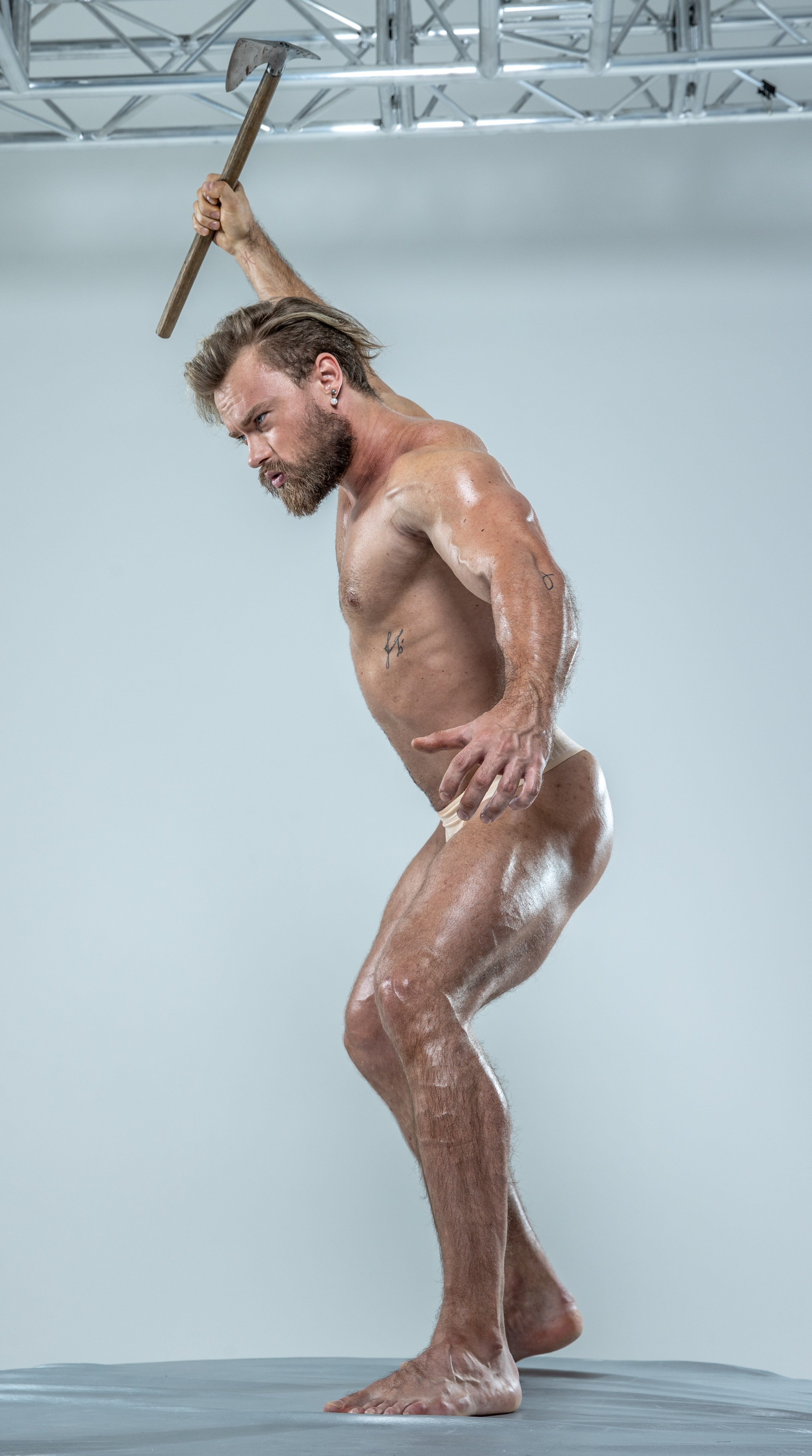 a muscular, bearded man with an axe raised above his head, captured in midaction against a minimalist studio backdrop. the image emphasizes his physique and the dynamic energy of the pose, highlighted by strategic lighting and a neutral background.