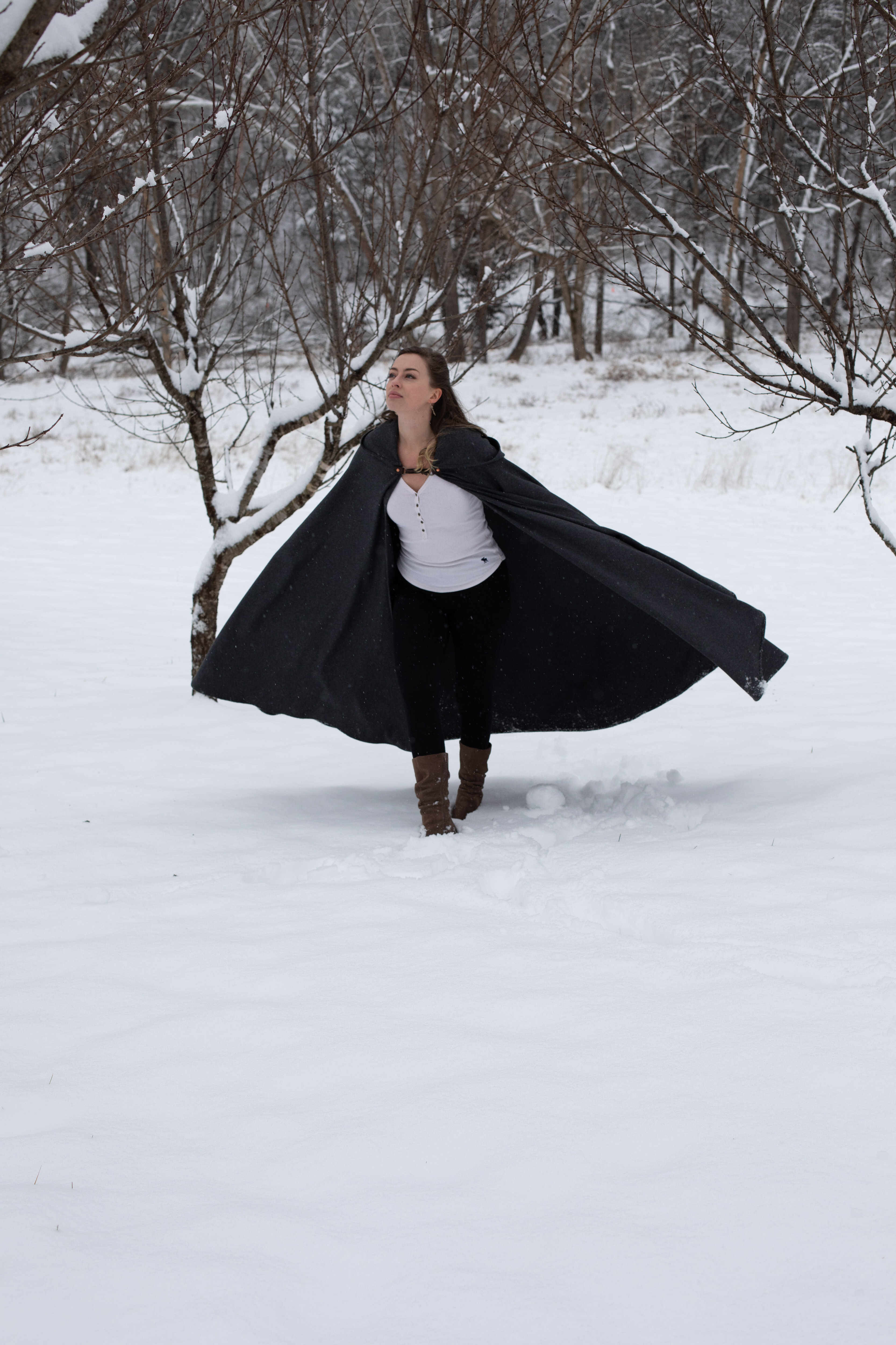 a young woman stands in a snowy forest, her arms outstretched beneath a flowing black cloak. the snowcovered trees and overcast sky create a serene and wintry atmosphere.