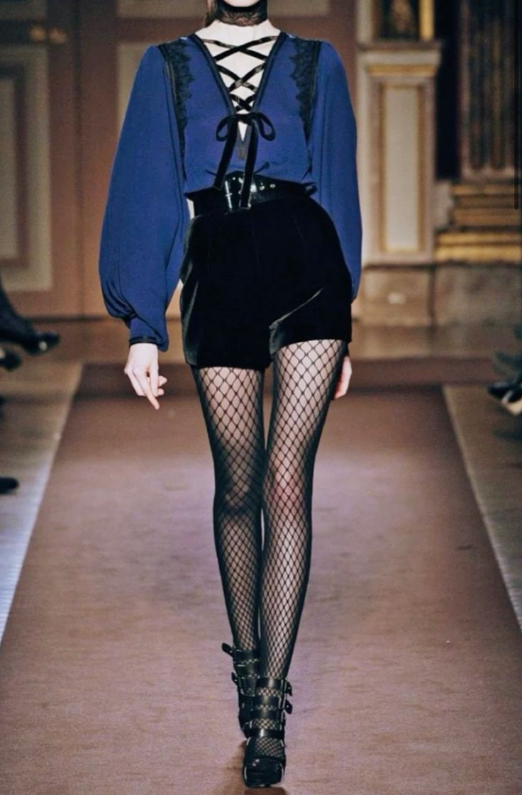 a female fashion model is walking down a runway wearing a blue shortsleeved blouse and black shorts. she is also wearing black fishnet stockings and black heeled boots with buckles. she looks elegant and stylish, showcasing a trendy outfit. the image has a professional fashion shoot feel.