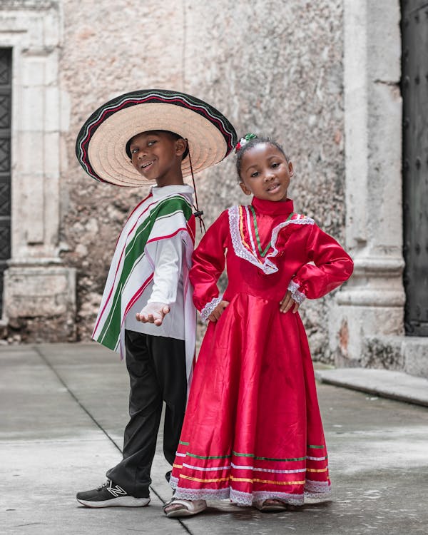 two young children, a boy and a girl, are dressed in vibrant traditional mexican attire. the boy wears a large sombrero and a green, white, and red ponchostyle shirt with dark pants. the girl is in a bright red dress with white lace and colorful stripes at the hem. they stand side by side, smiling, in front of an old stone building. the scene evokes a sense of cultural pride and tradition.