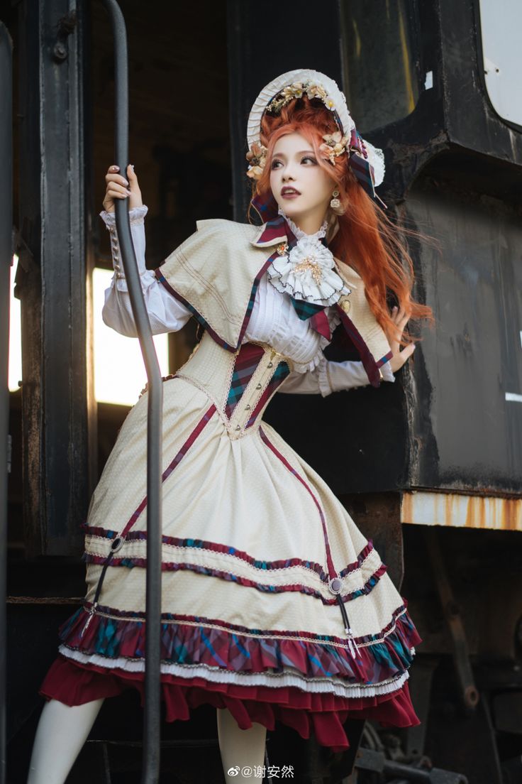 a young woman in a vintage inspired dress, corset, and hat poses beside a train car. she has long red hair and is holding onto the railing of the car as she looks away. the dress is white and cream with red and black plaid patterns. the train car is old and worn, with a rusty exterior. the image is in focus and well lit.