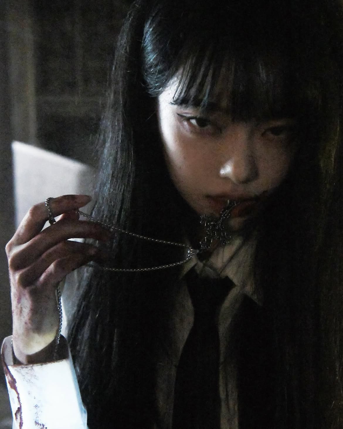 a dark, creepy image of a woman with pigtails and bangs, holding a necklace in her hand and biting on the pendant. she has dark makeup and is wearing a white shirt and a black tie. there appears to be blood on her hand and sleeve, adding to the horror aesthetic.