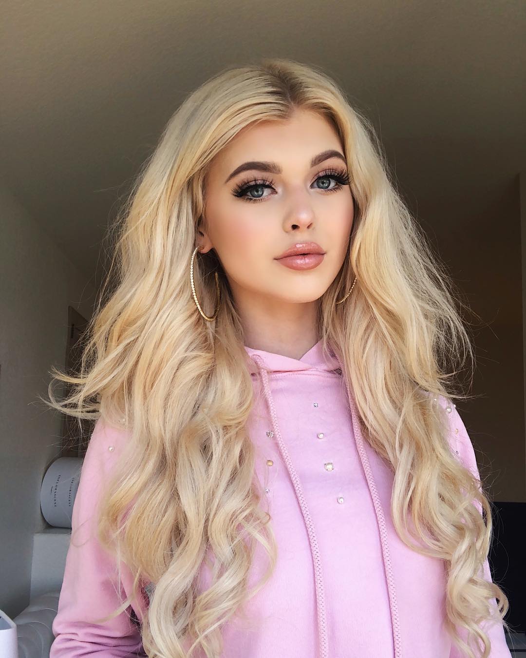 a portrait of loren gray, a young woman with long blonde wavy hair, wearing a pink hoodie with small gems, and large gold hoop earrings. she has fair skin, blue eyes, and is wearing makeup, including false eyelashes and lip gloss. she is posing indoors.