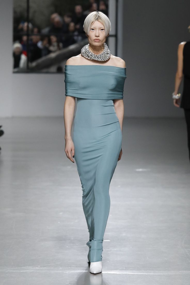 a female fashion model walks the runway in a light blue off the shoulder dress. she is wearing a large silver necklace and white high heels. her hair is styled in a short blonde bob. she is walking with a serious expression on her face. the background is blurred and shows other people in the audience.