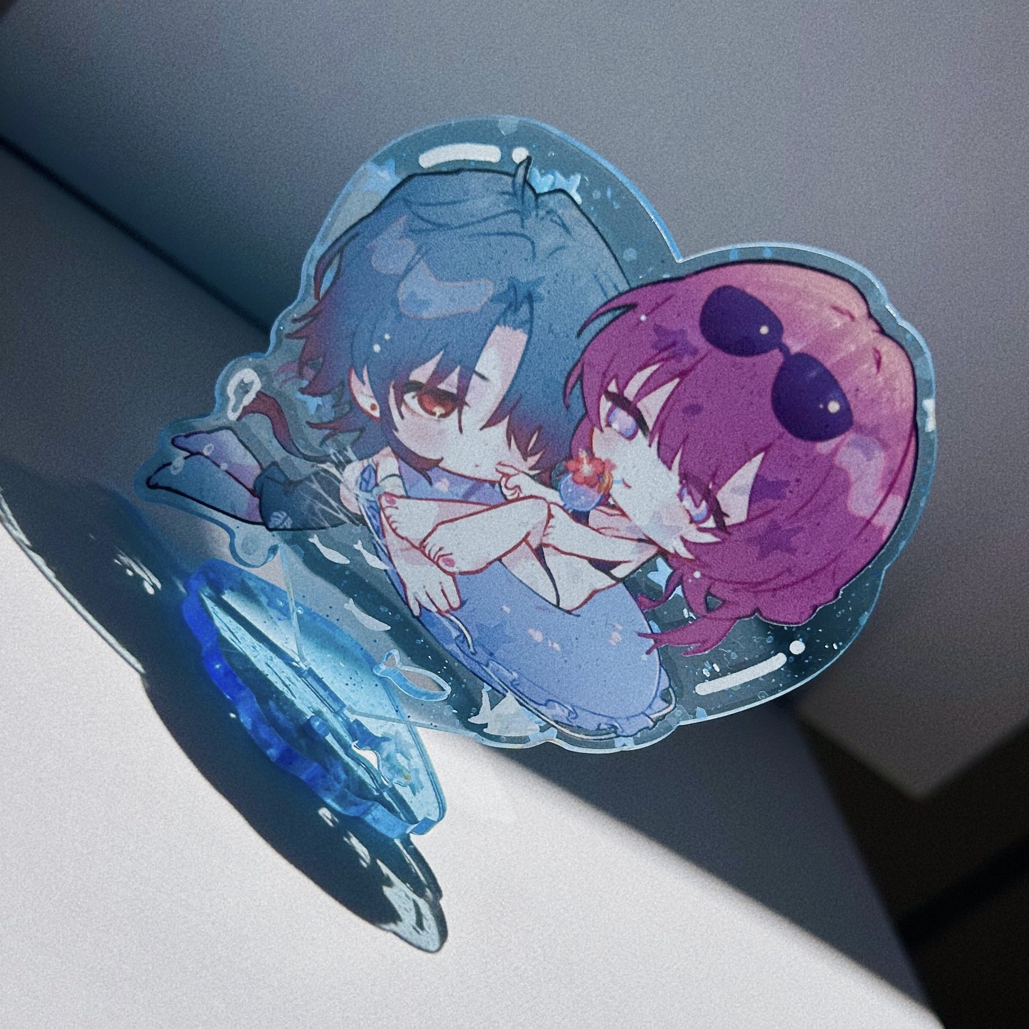 a closeup shot of an anime figurine featuring two characters, a boy and a girl, hugging. they are both in a cute chibi style and are surrounded by blue water. the boy has blue hair and is wearing a blue shirt, and the girl has pink hair and is wearing a purple and blue outfit. the figurine is transparent and features a stand to hold it up.  this could be a fanmade collectible or a commercially produced item.