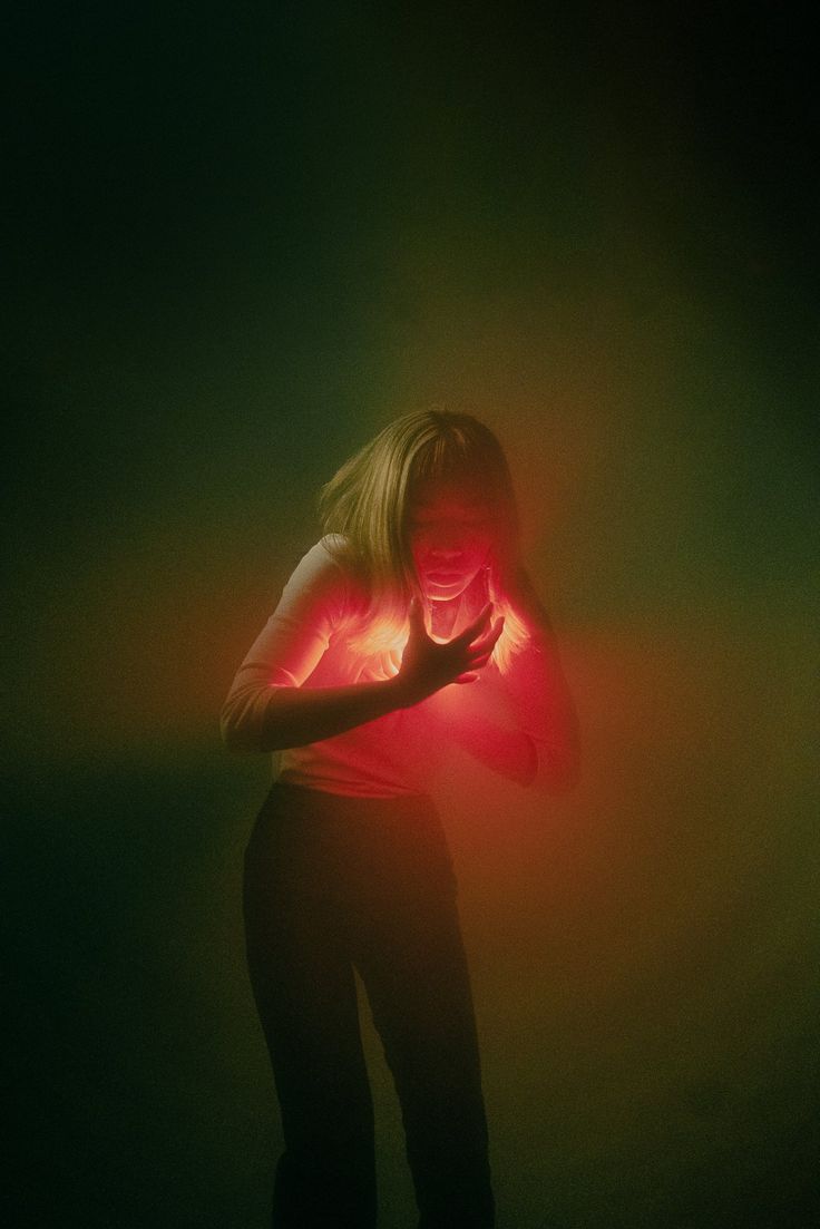 a woman stands in a dark studio lit with red light that casts a glow on her face and hands as she gestures to her heart. the background is a hazy green. her expression is intense and she looks like she is in pain or heartache.