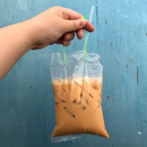 a person's hand is holding a clear plastic bag filled with iced teh tarik (a popular south east asian beverage). the bag has a straw inserted and is tied with green plastic ties. the background is a worn, tealcolored wall. the image suggests a street food or takeaway context.