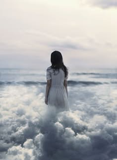 a woman wearing a white dress stands alone on a beach with clouds surrounding her, looking wistfully out towards the ocean.