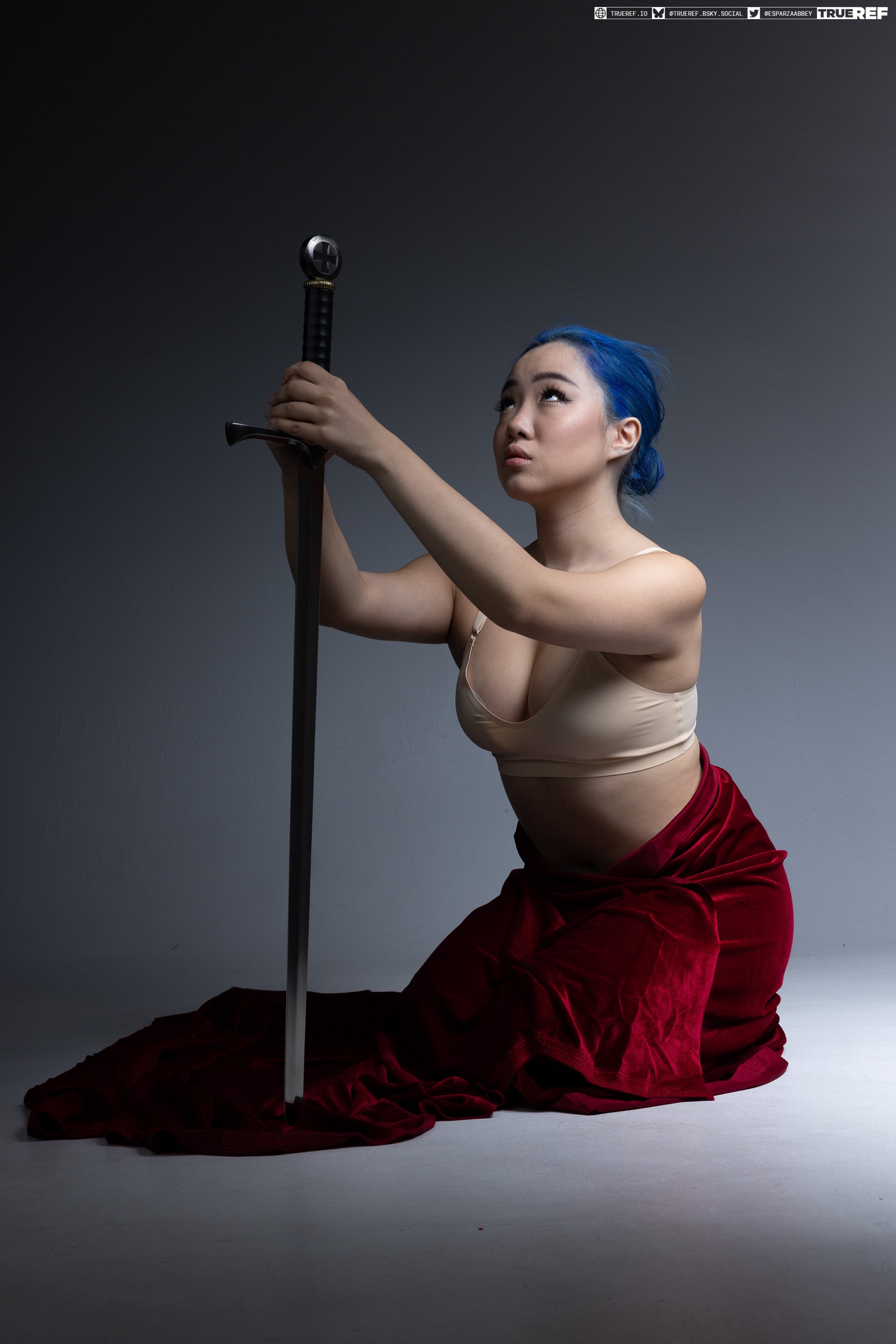 an asian woman with blue hair kneels in a studio setting while holding a medieval sword upright. she is wearing a tan bra and a red velvet skirt that pools around her. her gaze is directed upward.