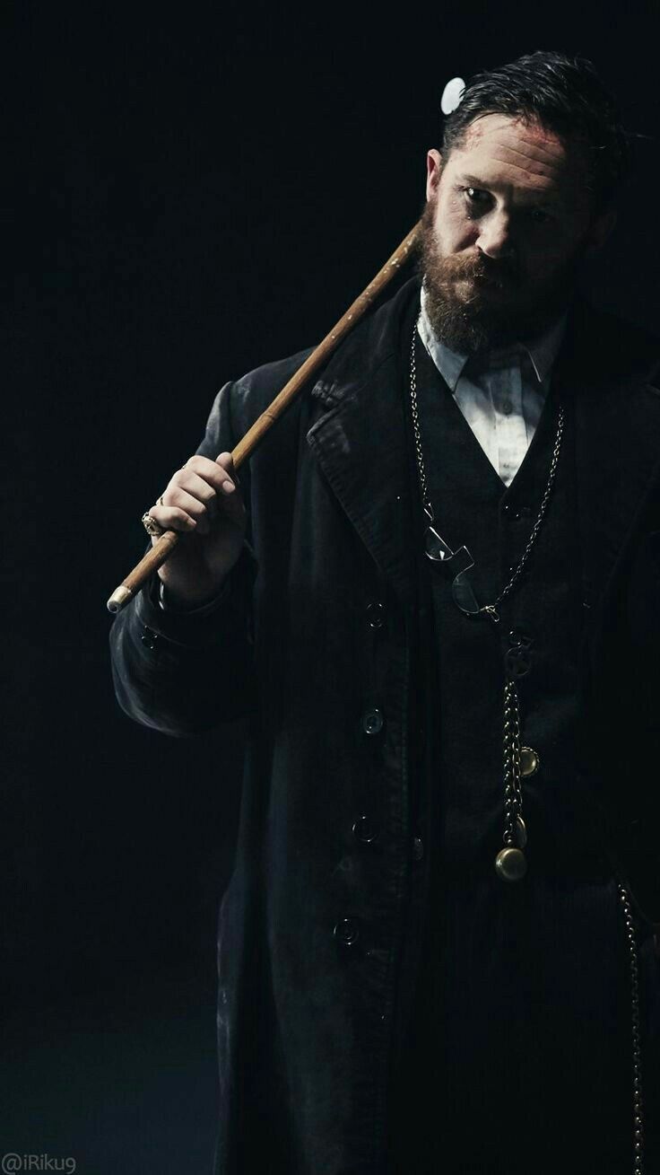 a portrait of tom hardy in character, likely as alfie solomons from peaky blinders. he is dressed in a dark coat over a vest, shirt, and tie. he holds a cane across his shoulders. a chain with glasses and a pocket watch hangs from his neck. the image is dark with dramatic lighting.
