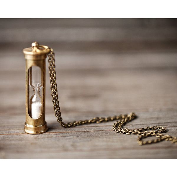 a gold chain necklace with a brass hourglass pendant sits on a wooden surface. the chain is made of a thin, delicate chain and the pendant is a small hourglass.  the background is blurred with a warm wooden tone.