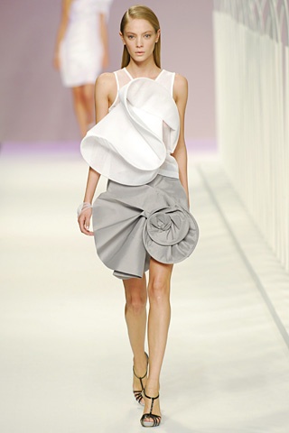a model wearing a white top and grey skirt walks the runway during a fashion show. the model is wearing black heels and looks elegant and stylish. the model has long blonde hair and natural makeup. the background is blurred and there are white railings in the distance.