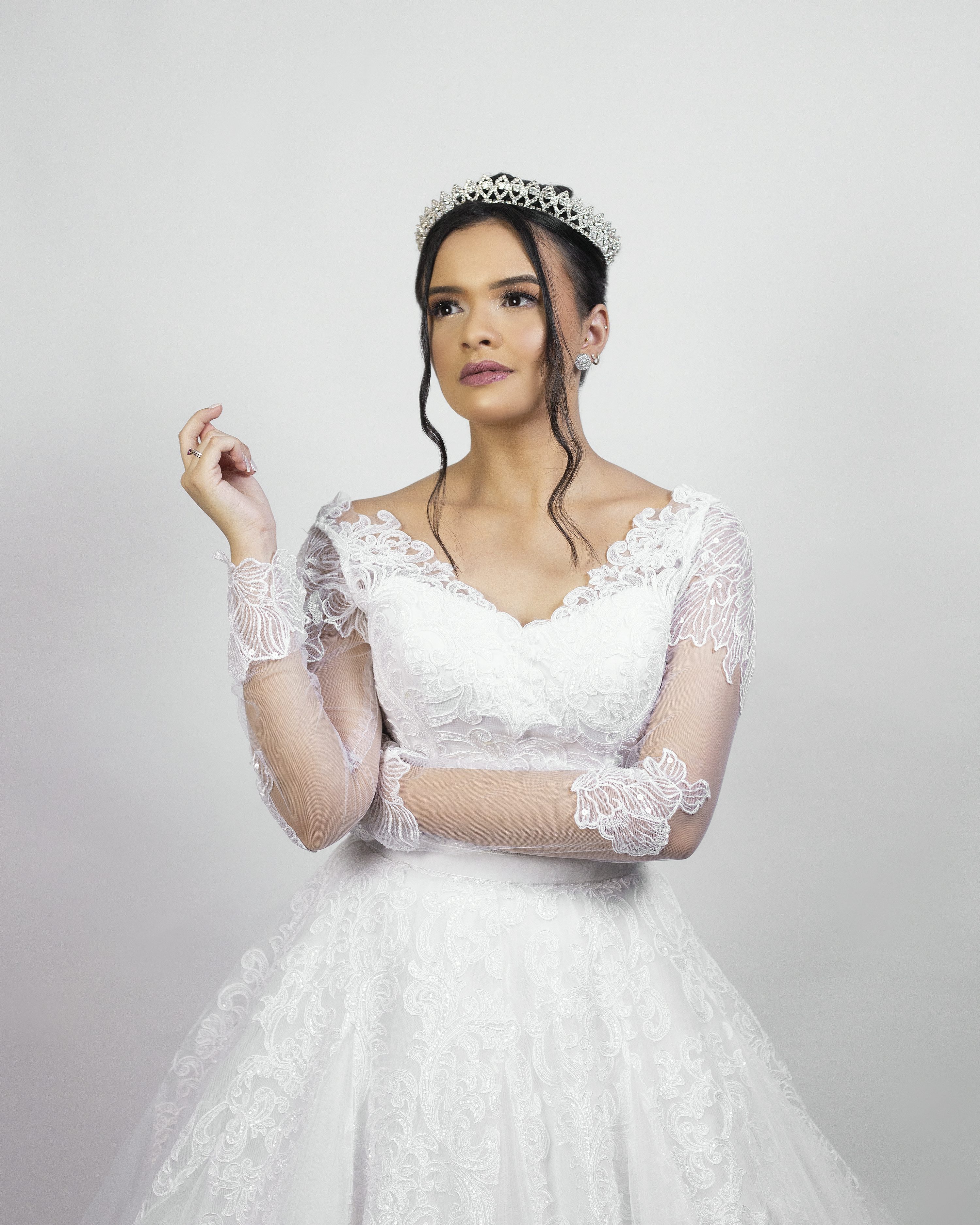 the image showcases a beautiful bride wearing a stunning white lace wedding gown with long sleeves. the dress has a fitted bodice and a full, flowing skirt. she is adorned with a sparkling tiara and her hair is styled in a way that complements the dress. the bride's makeup is subtle and elegant, enhancing her natural beauty. she gazes pensively off to the side, creating a serene and romantic atmosphere. the background is a simple, offwhite backdrop, allowing the dress and the bride to be the focal point of the image.  the overall feeling is one of classic bridal elegance.