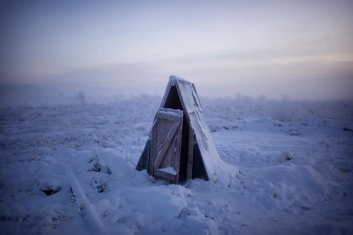 a small wooden shelter sits in a snow covered landscape. the snow is deep and white, and the sky is a soft blue. the shelter has a door that is slightly ajar, and the interior is dark. the image is serene and peaceful, and it evokes a sense of solitude and isolation. the winter landscape is a vast expanse of white snow, with the shelter in the foreground. the fog, which covers the ground in the background,  gives an ethereal feeling to the landscape.