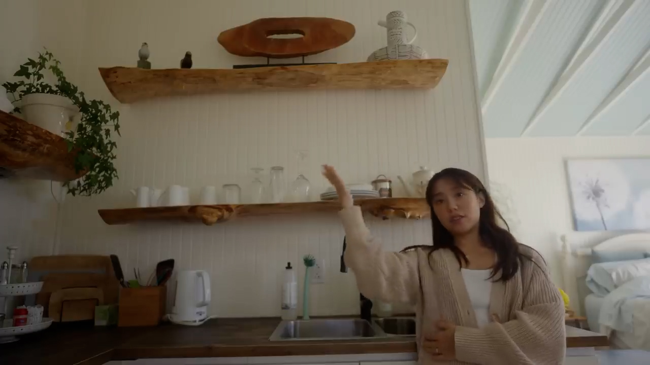 a woman stands in a kitchen with white cabinets and counters. above the counters are two liveedge wooden shelves.  the top shelf holds a piece of art and two vases. the bottom shelf holds glasses, a pitcher, and two canisters. to the left of the shelves is a small plant. behind the woman is a bedroom with a bed and a blue ceiling. the overall feeling of the space is bright, airy, and inviting.