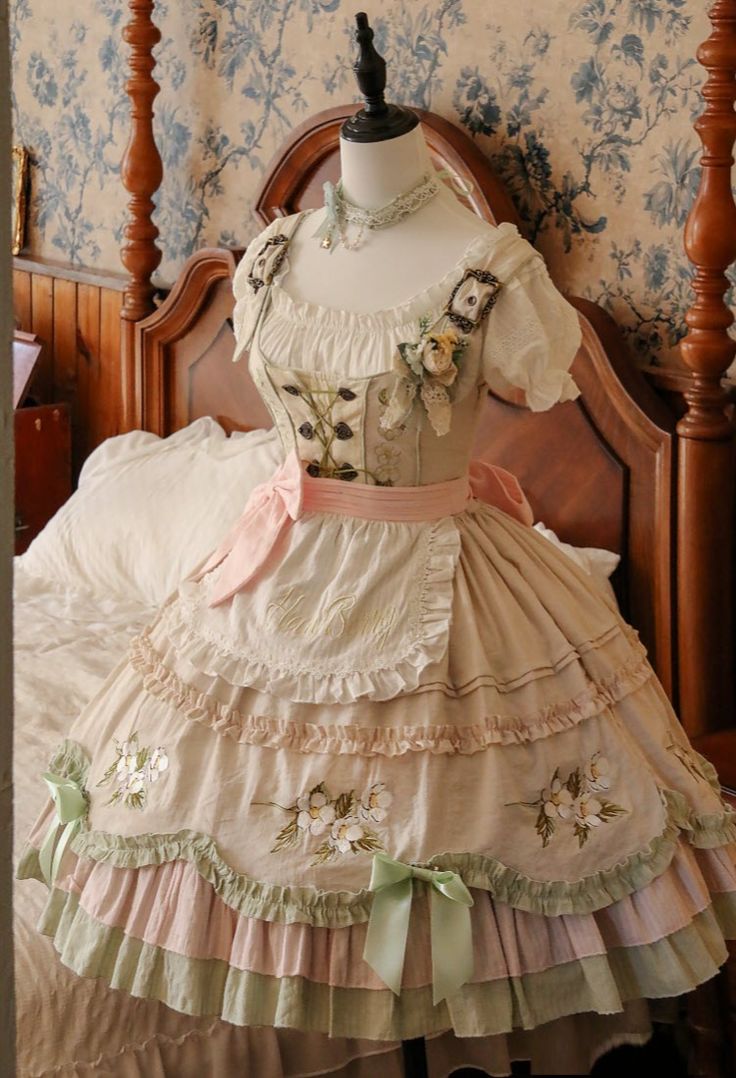the image showcases a detailed view of a "classic lolita" style dress draped over an antique bed.  the dress, crafted in pastel shades of cream, green, and pink, features intricate floral embroidery, lace trim, and a layered skirt with bows. the setting, an antique bedroom with ornate woodwork and floral wallpaper, further emphasizes the vintage aesthetic.
