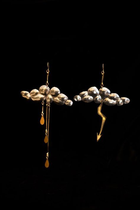 the image shows a pair of earrings in a dark setting. one earring features a cloudshaped design with gold rain droplets hanging below, while the other earring has a cloud with a gold lightning bolt.