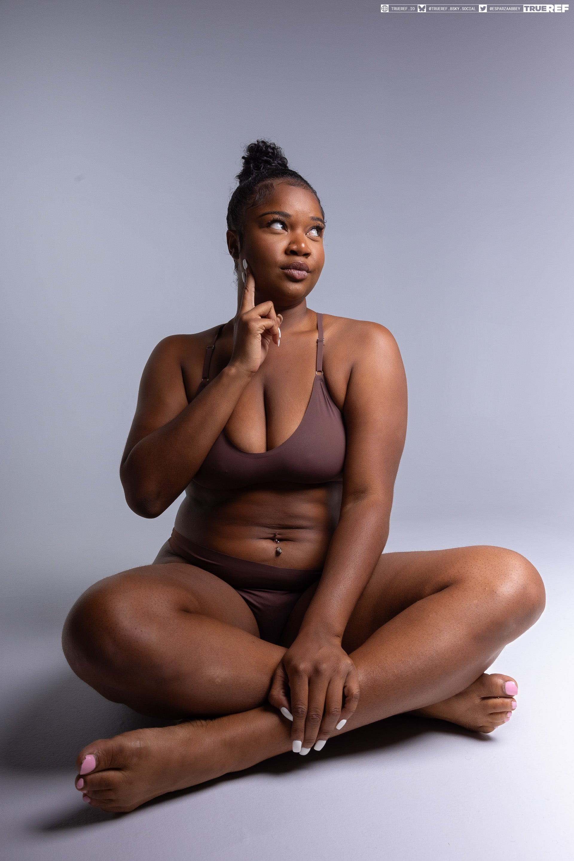 a woman with dark skin is sitting crosslegged in a studio shot. she is wearing brown lingerie and has her index finger pointing to her temple, as if she is thinking. her belly button is pierced and her fingers and toes are painted with nail polish.