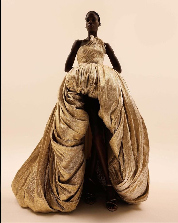 a fulllength studio shot features a black woman standing confidently while posing in a gold, oneshoulder gown. the dress is heavily draped, creating a voluminous and dramatic silhouette. she is wearing strappy sandals. the background is a neutral, offwhite color.
