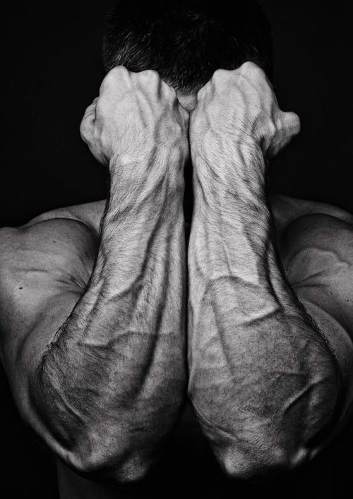 the image showcases the back of a muscular man's upper body. his arms are bent, and his hands are clasped behind his head, highlighting the definition and veins in his forearms, biceps, and back. the photo is taken in black and white, emphasizing the textures and contours of his physique. the background is dark, directing all focus to the man's body.