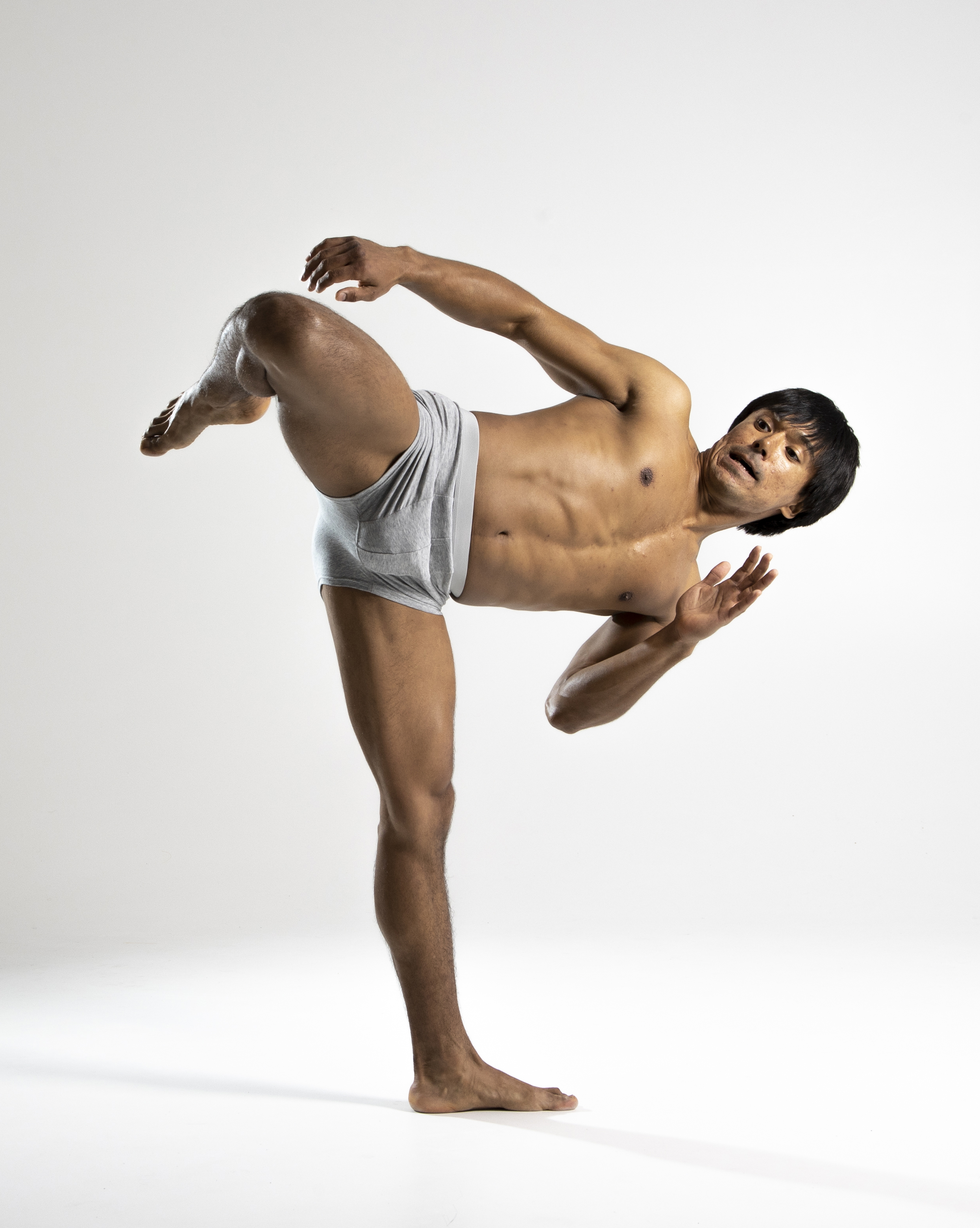 a shirtless athletic man in gray underwear is captured in a dynamic pose against a white background. he is standing on his right leg while kicking his left leg up towards his body. his body is turned slightly to the right, with his arms outstretched and his facial expression conveying intensity. he appears to be a dancer or someone engaged in a fitness activity that requires flexibility and balance.