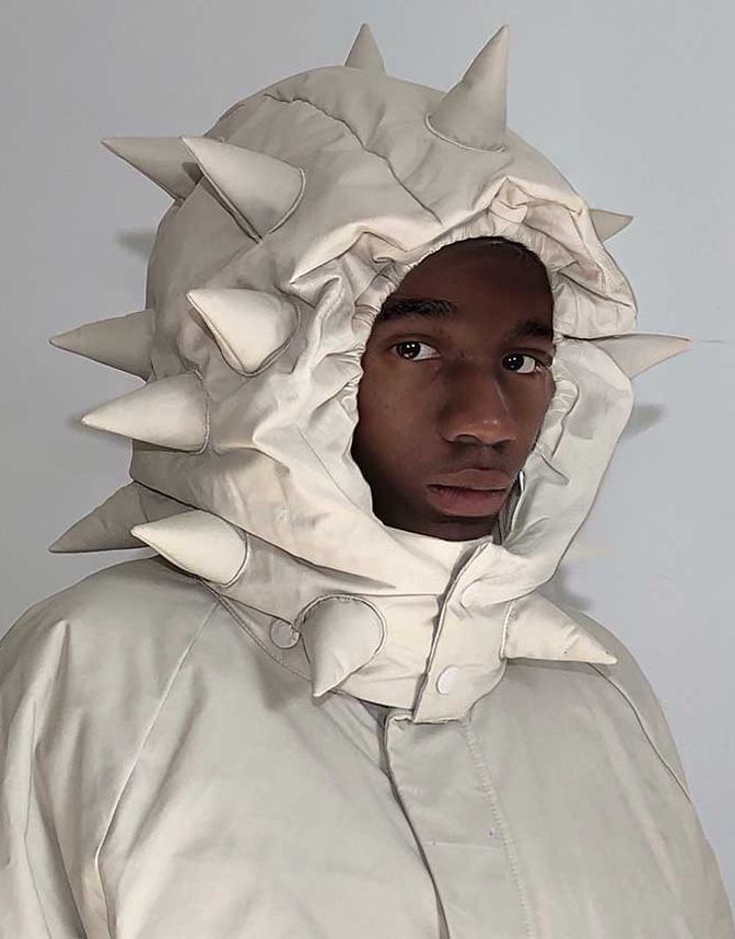 a man is wearing a white puffer jacket with a large hood covered in spikes. the jacket is unzipped and the man is looking directly at the camera.