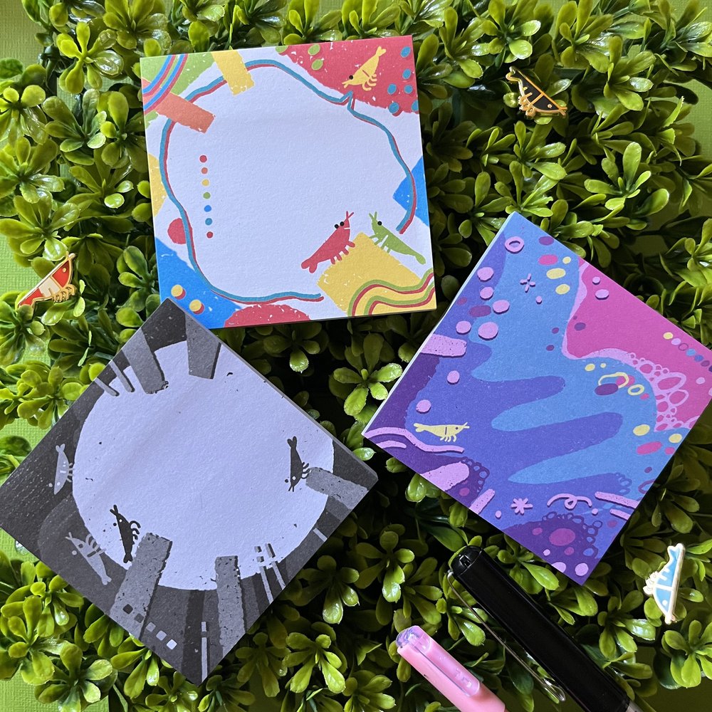 the image shows three square sticky notepads arranged on a bed of green boxwood.  each notepad features a unique abstract design incorporating small illustrations of shrimps or prawns in different color palettes.  the pads have a soft, pastel color scheme, with one being predominantly purple and pink, another predominantly warm tones, and a third using grey and black.  a black pen and a pink gel pen are also in the image, adding to the stationery theme.  the overall feel is one of playful, artistic design and organization.
