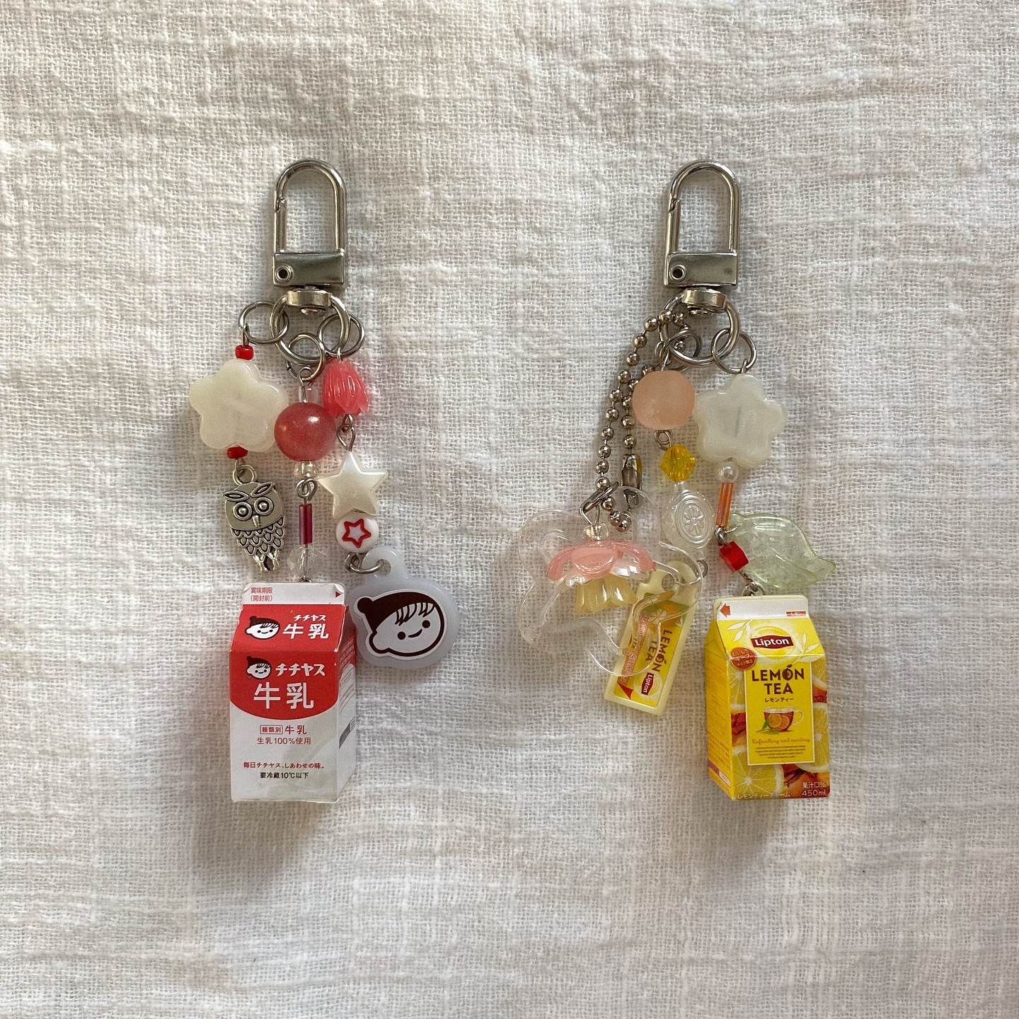 the image shows two handmade keychains on a white textured fabric. one keychain features a miniature red and white japanese milk carton, an owl charm, white star, and pink beads. the other keychain has a small yellow lipton lemon tea box, a yellow candy charm, a pink jellyfish charm, green leaf charm, and various beads. the keychains both have silver clasps and chains.
