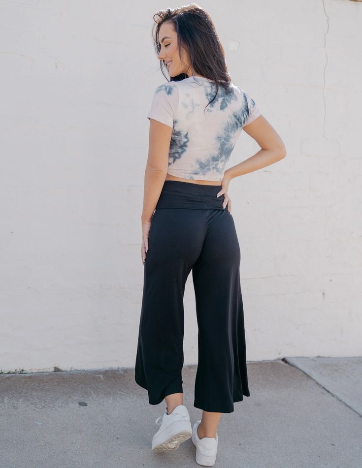 a young woman with long brown hair stands with her back to the camera, showcasing a stylish casual outfit. she wears a cropped tiedye tshirt in shades of white and grey, paired with black wideleg pants that have a folded waistband. her white sneakers complement the overall relaxed yet chic look. the setting appears to be an urban environment with a white textured wall in the background, and the natural lighting illuminates the scene brightly.