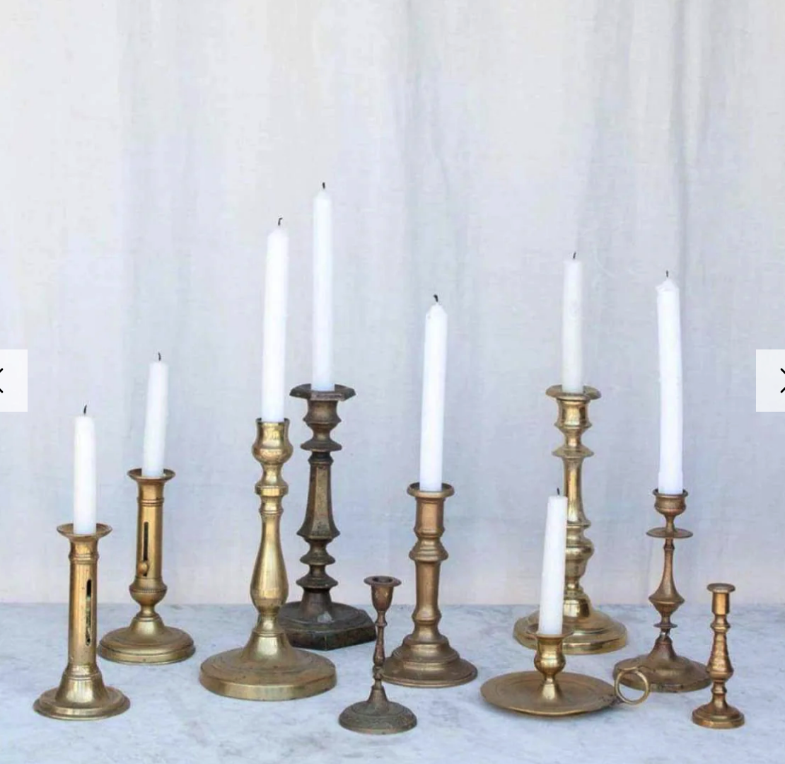 a collection of brass candlesticks of different sizes and designs, with white candles in them, set on a white surface.  they would make a beautiful centerpiece for a table.