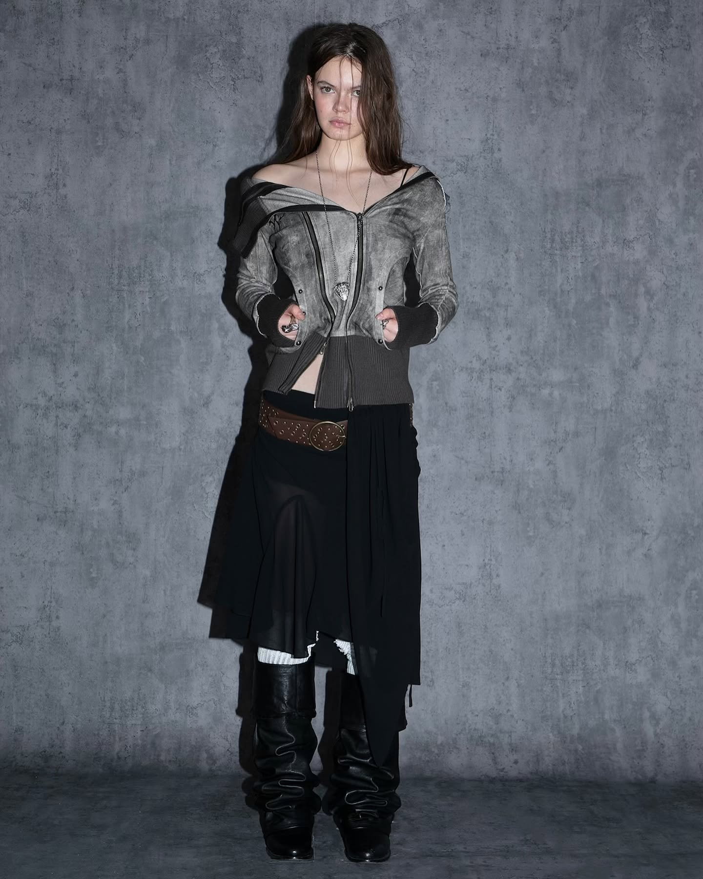 a fulllength studio shot of a young woman modeling a dark and edgy outfit. she is wearing a grey and black distressed jacket with a partially unzipped front.  the jacket has a slightly offtheshoulder style.  she's also wearing a black layered skirt that appears to be partially sheer and has an asymmetrical hemline. brown studded belt is visible at her waistline. black slouchy boots complete her look.  the background is a textured grey wall. the overall style is reminiscent of grunge, gothic, or alternative fashion. the model has long brown hair and neutral makeup. she's looking directly at the camera.