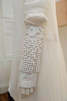a closeup view of the detail on a white gown. the sleeve is covered in a delicate beaded pattern that gives the dress a luxurious and elegant look. the fabric is white, and it's hard to tell if the beads are sewn on or if they are a part of the fabric itself. the dress is made with high quality materials and craftsmanship, making it a unique piece of fashion.